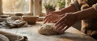Elderly hands kneading rustic bread dough on a flour-dusted wooden table in a warm, sunlit farmhouse kitchen with bowls nearby.