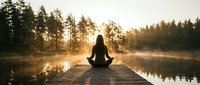 Woman practicing yoga meditation on wooden dock at sunrise over misty lake surrounded by forest trees creating peaceful wellness retreat atmosphere.