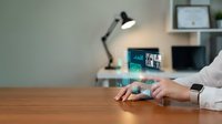 Person wearing white long-sleeve shirt and smartwatch using holographic digital interface displaying financial charts and data visualization on wooden desk with desk lamp and shelving.
