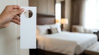 Close-up of hand holding blank white door hanger tag in modern hotel bedroom with bed and furniture visible in soft focus background for hospitality marketing.