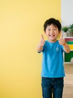 Young Asian boy, approximately 5-7 years old, wearing blue t-shirt and jeans, giving double thumbs up gesture with bright smile against yellow wall background. Suitable for education, parenting, success and positivity concepts.