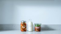 Fresh organic vegetables preserved in glass jars with milk bottle on clean white surface for healthy meal prep and sustainable food storage.
