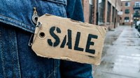 Person in denim jacket holding a cardboard sale tag on a city street, conveying discount, retail promotion, and urban shopping concept.