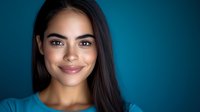 Young Hispanic woman with long dark hair and radiant smile against blue background, professional headshot style portrait showcasing natural beauty and confidence.