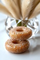 Sugar-coated mini donuts stacked on white surface with crystal vase and dried pampas grass in soft focus background, creating elegant composition.