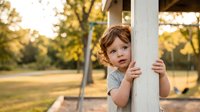 Young toddler girl with curly brown hair peeking around white wooden post on playground during golden hour sunset with warm natural lighting and bokeh background.
