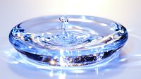 Mesmerizing water drop impact creating crown splash formation in clear liquid with blue tint, captured in high speed photography on reflective surface.