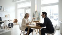 Professional blonde businesswoman and bearded businessman having meeting at wooden table in bright modern office space with large windows and minimalist decor.