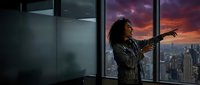 Woman with curly dark hair, wearing a patterned jacket, standing by floor-to-ceiling windows in a high-rise office, pointing toward a dramatic sunset cityscape below.