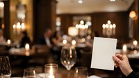 Waiter holding blank menu card in upscale restaurant with elegant table setting, candlelight, and guests dining in background for hospitality and service concepts.