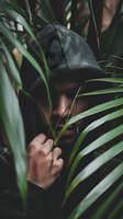 Mysterious hooded figure partially hidden behind tropical palm leaves in moody atmospheric lighting, creating dramatic contrast between shadow and foliage.