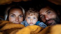 Happy family with young child snuggled under warm yellow blanket, close-up portrait showing bright blue eyes and intimate connection between parents and toddler son.
