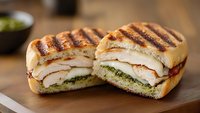 Grilled panini sandwich cut in half showing layers of turkey, cheese and pesto on wooden cutting board. Perfect for restaurant menus and food blogs.