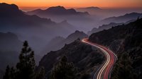 Winding mountain road with light trails at sunset creating dramatic orange and purple sky over layered mountain ridges in scenic landscape.