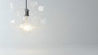 Glowing light bulb with business icons floating around it representing innovation, creativity and bright ideas in modern minimalist interior space.