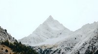 Snow-capped mountain peak rises above evergreen forest and rocky terrain in dramatic winter landscape, captured in muted tones against cloudy sky.