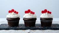 Three chocolate cupcakes with white frosting topped with red heart decorations and colorful sprinkles on dark slate board for Valentine's Day celebration.