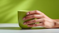 Female hands with natural manicure holding green ceramic mug on green background. Minimalist composition with soft shadows for wellness and lifestyle design.