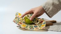 Caucasian hand wrapping fresh green apple in reusable beeswax wrap with floral pattern on wooden board, eco-friendly zero waste kitchen concept.