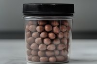 Chocolate covered almonds in clear plastic jar with black lid on gray background, selective focus on nuts creating depth and texture in minimalist composition.