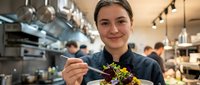 Young woman with light skin tone and brown hair, wearing dark chef's coat, plating gourmet dish with microgreens in professional kitchen. Culinary professional demonstrating food preparation and fine dining expertise.