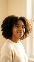 Young African American woman with natural curly hair smiling warmly in cream sweater by window with soft golden light for lifestyle and wellness content.