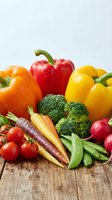 Fresh organic vegetables including red, orange and yellow bell peppers, broccoli, carrots, tomatoes, radishes and snap peas arranged on wooden surface for healthy eating and nutrition.