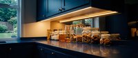 Modern kitchen countertop with warm under-cabinet lighting, dark cabinetry, and organized storage containers. Suitable for home design, interior decoration, and lifestyle publications.