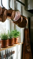 Rustic kitchen windowsill with copper pots hanging on rack, fresh herb plants in terracotta pots, and leather apron for cozy farmhouse cooking decor.
