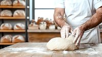 Male baker kneading fresh dough on floured wooden surface in bakery, wearing white uniform and gloves, with artisan bread loaves on shelves in background.