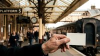 Senior man holding blank ticket at railway station platform with vintage train and passengers in background, suitable for travel, transportation, and nostalgia concepts.