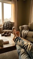 Man relaxing at home with bare feet up on rustic wooden coffee table, cozy living room with warm sunlight, jeans, book, mug, and leather shoes nearby.