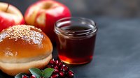 Fresh burger bun with sesame seeds alongside red apples, pomegranate seeds and honey or maple syrup in glass jar on dark background, food styling.