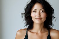 Young Asian woman with natural makeup and tousled black hair smiling confidently against light background, expressing genuine joy and optimism in casual portrait.