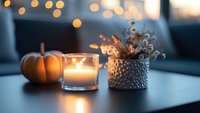 Cozy autumn evening with glowing candle, decorative pumpkin and dried flowers in textured vase against blurred bokeh lights creating warm atmospheric mood.
