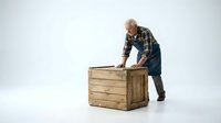 Senior Caucasian craftsman in plaid shirt and apron examining handmade wooden crate on white background. Woodworking artisan quality control concept.