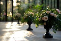 Elegant floral arrangement with blush roses and mixed greenery in classic black urn, backlit by soft natural sunlight creating romantic atmosphere at sunset.