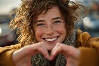Young woman with curly hair making heart shape with hands, smiling joyfully outdoors in warm sunlight, expressing love and happiness.