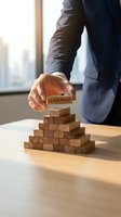 Businessman in navy suit placing wooden block labeled leadership on pyramid stack at office desk with city skyline window background, representing business growth and management strategy.