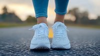 Close up of white running shoes on asphalt road during sunset workout. Athletic footwear and blue leggings shown from front view against blurred nature background.
