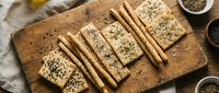 Sliced seeded bread and whole grain crackers arranged on wooden cutting board with sesame and poppy seeds for baking and cooking projects.