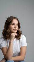 Young Caucasian woman with wavy brown hair in white t-shirt sitting thoughtfully with hand on chin against gray background for lifestyle content.