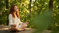 Young Caucasian woman with curly red hair meditating in lotus pose on wooden deck surrounded by lush green forest in sunlight.
