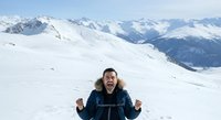 Young man with dark hair and light skin, wearing a dark blue winter jacket with fur-lined hood, expressing excitement and joy while sledding down snowy mountain slopes with alpine peaks in background.
