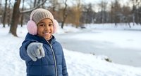 Happy African American girl in winter coat and knit hat pointing at camera in snowy park. Child enjoying outdoor winter activities and seasonal fun.