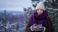 Young Asian woman in winter clothing holding snow in cupped hands outdoors with evergreen trees and mountain landscape background during twilight hour.