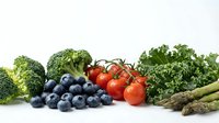 Fresh organic vegetables and berries including broccoli, kale, cherry tomatoes, blueberries and asparagus arranged on white background for healthy eating.