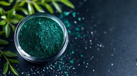 Vibrant blue-green spirulina powder in glass jar with scattered powder and fresh bamboo leaves on dark background, top view composition for wellness and nutrition.