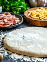 Fresh pizza dough on dark background with prosciutto and cheese ingredients, ready for baking. Homemade Italian cuisine preparation with flour dusted surface.