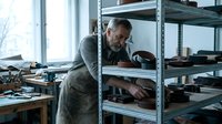 Mature male craftsman with gray beard and dark hair, wearing work apron, organizing tools and materials on metal shelving in bright workshop studio space.